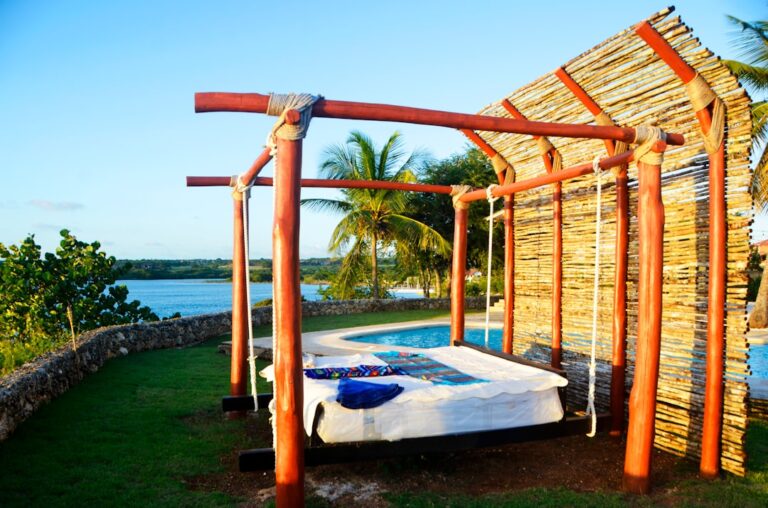 Uma Cama Sentada Sob Uma Estrutura De Madeira Ao Lado De Um Corpo De Agua v1yufeSLYiw