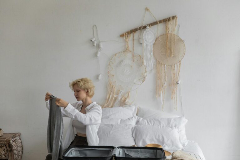 Tranquil female with blond hair sitting on bed and putting garment in suitcase while preparing for travel in cozy room