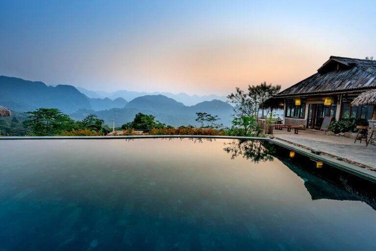 Pool with calm water located near terrace of tropical resort with thatched house against mountain ridge and green trees in nature